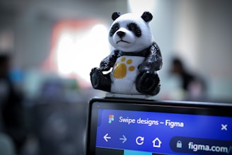 A small panda figurine with a yellow paw print sits atop a computer screen displaying the Figma website. The background is blurred, likely indicating an office setting.