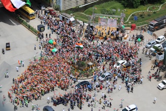 A large crowd gathers at an intersection for a public event. Many people hold flags and banners featuring the colors of the Indian national flag, creating a sea of orange, white, and green. In the center, there is a small roundabout with greenery, and a stage is set up with people on it. Vehicles, including buses and cars, are parked on the road. The atmosphere appears festive and organized.