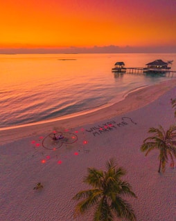A serene beach scene at sunset with a vibrant orange and pink sky. Two overwater bungalows are visible in the background. On the sandy shore, there is a romantic setup with a heart shape outlined in red lights and a couple seated inside. The phrase 'WILL YOU MARRY ME?' is written in the sand.