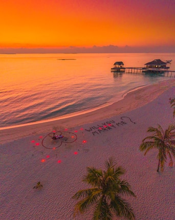 A serene beach scene at sunset with a vibrant orange and pink sky. Two overwater bungalows are visible in the background. On the sandy shore, there is a romantic setup with a heart shape outlined in red lights and a couple seated inside. The phrase 'WILL YOU MARRY ME?' is written in the sand.