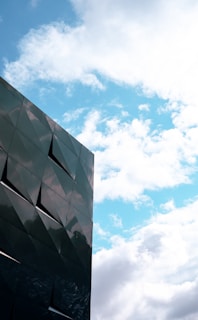 A modern building with a facade of dark, reflective panels is set against a bright blue sky scattered with white clouds.