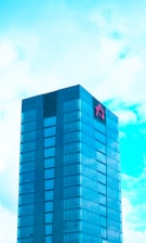 A modern glass skyscraper with a blue tint stands tall against a partly cloudy sky. The building features reflective windows and a prominent red logo near the top.
