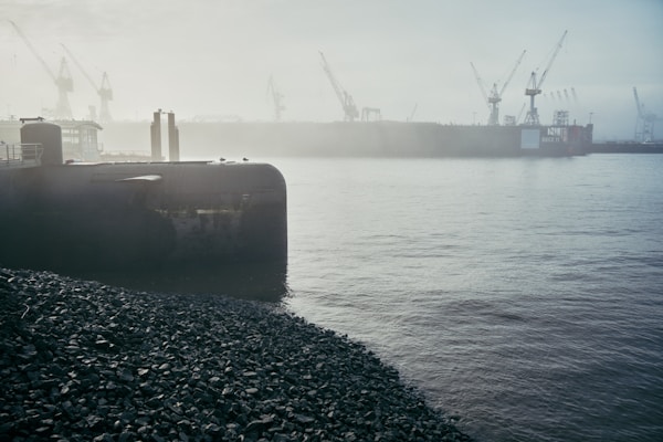 A large submarine is partially submerged in the water, positioned next to a rocky shoreline. Several cranes are visible in the background, partially obscured by fog, suggesting an industrial or shipyard setting. The atmosphere is misty, with the platform giving a stark contrast to the calm waters.