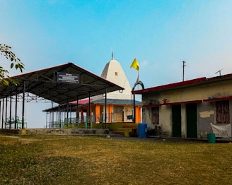 A small temple with a domed roof and a yellow flag on top is situated in an open area. To the left, there is a covered structure with green benches beneath a metal roof. The ground is grassy, and the sky is clear and blue. A wall bears the name of the temple.