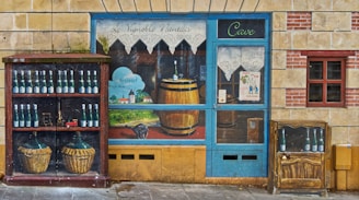 A mural depicts a vineyard and a storefront scene painted on a wall. The artwork includes a variety of wine bottles, a wooden barrel, and two wicker baskets. The image features a blue door with lace curtains, a poster advertising a jazz event, and a detailed landscape with a small village and hills. The overall aesthetic combines rustic and artistic elements, creating an illusion of a wine shop facade.
