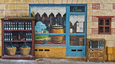 A mural depicts a vineyard and a storefront scene painted on a wall. The artwork includes a variety of wine bottles, a wooden barrel, and two wicker baskets. The image features a blue door with lace curtains, a poster advertising a jazz event, and a detailed landscape with a small village and hills. The overall aesthetic combines rustic and artistic elements, creating an illusion of a wine shop facade.