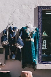Two traditional garments are displayed against a white textured wall. One is a blue robe with white stripes and wooden buttons, and the other is a teal dress with intricate embroidery and gold detailing. They are hung on hangers, and a photo frame with a geometric patterned border is visible nearby.
