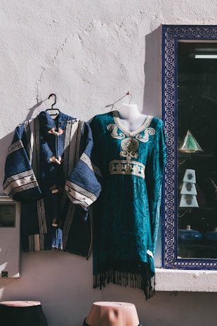 Two traditional garments are displayed against a white textured wall. One is a blue robe with white stripes and wooden buttons, and the other is a teal dress with intricate embroidery and gold detailing. They are hung on hangers, and a photo frame with a geometric patterned border is visible nearby.