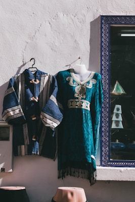 Two traditional garments are displayed against a white textured wall. One is a blue robe with white stripes and wooden buttons, and the other is a teal dress with intricate embroidery and gold detailing. They are hung on hangers, and a photo frame with a geometric patterned border is visible nearby.