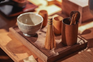 A wooden tray holds a ceramic bowl, a bamboo whisk, and a cylindrical container containing wooden utensils. The setting is warmly lit, creating an inviting and rustic ambiance, with the natural wood grains and earthenware tones adding to the aesthetic.