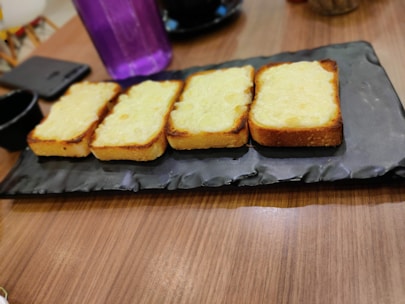 Four slices of toasted bread topped with melted cheese are arranged on a rectangular textured black platter. The background includes a blurred view of a purple water bottle and other dining elements on a wooden table.