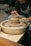 A person is working on a pottery wheel, shaping a clay vessel with skillful hands. The setting includes a stack of finished pottery pieces in the background, and the artist is focused on molding the clay.