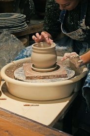 A person is working on a pottery wheel, shaping a clay vessel with skillful hands. The setting includes a stack of finished pottery pieces in the background, and the artist is focused on molding the clay.