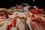 A selection of various fish and seafood arranged on a bed of ice, displayed at a market. Different types, shapes, and sizes of fish are visible with their scales and skin shining under the market lights. The assortment includes both whole fish and pieces cut into fillets, featuring vibrant colors and textures.