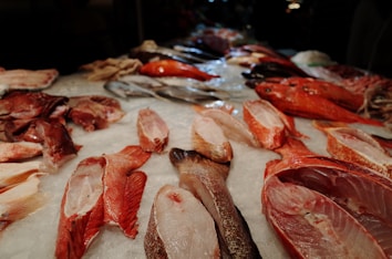 A selection of various fish and seafood arranged on a bed of ice, displayed at a market. Different types, shapes, and sizes of fish are visible with their scales and skin shining under the market lights. The assortment includes both whole fish and pieces cut into fillets, featuring vibrant colors and textures.