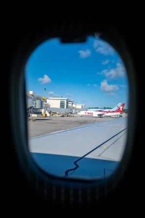 The view through an airplane window reveals an airport scene with an aircraft bearing the logo and colors of T'way Airlines parked at the terminal. Surrounding the plane, the tarmac is busy with airport equipment and vehicles. The sky is a clear blue with a few scattered clouds.