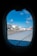 The view through an airplane window reveals an airport scene with an aircraft bearing the logo and colors of T'way Airlines parked at the terminal. Surrounding the plane, the tarmac is busy with airport equipment and vehicles. The sky is a clear blue with a few scattered clouds.