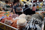 A busy market scene featuring various dried seafood products including small fish, shrimp, and squid. In the foreground, there are bags and piles of these items with clear, bright packaging. Several people can be seen in the background, some standing and others sitting at tables, all seemingly engaged in activities such as conversing or processing food. The market is well-lit with multiple light sources visible.