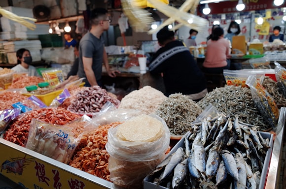 A busy market scene featuring various dried seafood products including small fish, shrimp, and squid. In the foreground, there are bags and piles of these items with clear, bright packaging. Several people can be seen in the background, some standing and others sitting at tables, all seemingly engaged in activities such as conversing or processing food. The market is well-lit with multiple light sources visible.