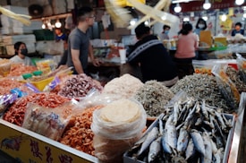 A busy market scene featuring various dried seafood products including small fish, shrimp, and squid. In the foreground, there are bags and piles of these items with clear, bright packaging. Several people can be seen in the background, some standing and others sitting at tables, all seemingly engaged in activities such as conversing or processing food. The market is well-lit with multiple light sources visible.