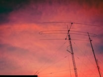 Silhouetted antennas stand against a vibrant sunset sky with hues of orange, pink, and purple. The antennas' intricate network of wires is clearly visible.