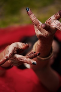 Intricately decorated hands with henna patterns pose against a blurred red and green background. The mehndi designs cover the fingers and palms, and the nails are painted black. A gold bracelet adorns one wrist, adding an elegant accessory to the traditional artwork.