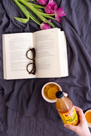 An open book with text visible lies on a dark fabric surface, accompanied by a pair of eyeglasses resting on its pages. A bouquet of pink tulips with green stems lies above the book. A hand is holding a bottle of apple cider vinegar, labeled in red and yellow, in the foreground. Next to it, a mug containing a drink with lemon slices is placed.