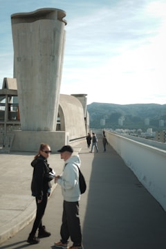 A large, modernist structure with a unique, artistic design features prominently. There are several people walking and interacting on a wide concrete surface, against a backdrop of distant hills and a clear sky.