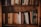 A bookshelf filled with a variety of hardcover books arranged closely together. The books have aged, neutral-colored spines, with some featuring gold or embossed lettering. The bookshelf is made of dark, polished wood, and the lighting is warm, giving the setting a cozy and nostalgic feel.