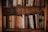 A bookshelf filled with a variety of hardcover books arranged closely together. The books have aged, neutral-colored spines, with some featuring gold or embossed lettering. The bookshelf is made of dark, polished wood, and the lighting is warm, giving the setting a cozy and nostalgic feel.