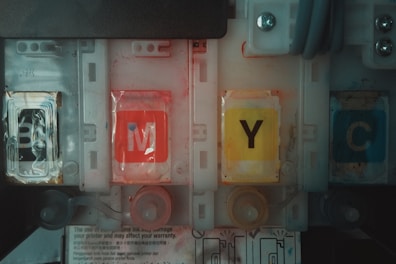 Four printer ink cartridges are aligned, labeled with the letters B, M, Y, and C. Each cartridge displays a distinct color: black, magenta, yellow, and cyan. The design includes plastic casing and visible internal compartments. The environment appears mechanical or industrial with hints of wear and smudges on the cartridges.