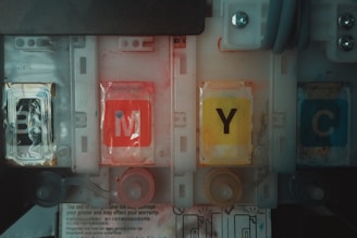 Four printer ink cartridges are aligned, labeled with the letters B, M, Y, and C. Each cartridge displays a distinct color: black, magenta, yellow, and cyan. The design includes plastic casing and visible internal compartments. The environment appears mechanical or industrial with hints of wear and smudges on the cartridges.