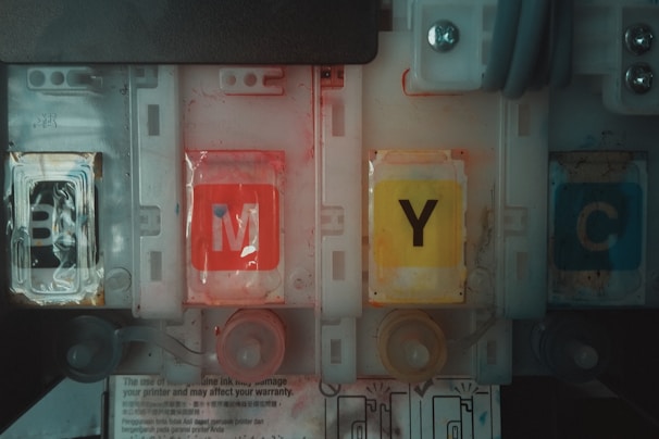Four printer ink cartridges are aligned, labeled with the letters B, M, Y, and C. Each cartridge displays a distinct color: black, magenta, yellow, and cyan. The design includes plastic casing and visible internal compartments. The environment appears mechanical or industrial with hints of wear and smudges on the cartridges.