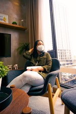 A person wearing a face mask and holding a tablet is seated in a modern room. The room features a large window with a cityscape view, a gray chair, a wooden table, and decorative plants. There is artwork and a small sculpture on the shelf.