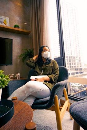 A person wearing a face mask and holding a tablet is seated in a modern room. The room features a large window with a cityscape view, a gray chair, a wooden table, and decorative plants. There is artwork and a small sculpture on the shelf.
