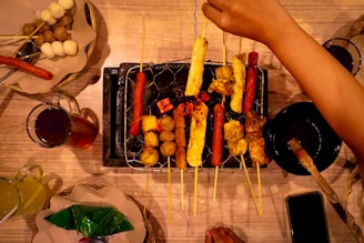 A variety of skewered foods are being grilled on a portable charcoal grill set on a wooden table. The skewers include sausages, meats, and possibly tofu or vegetables, with a hand adjusting them. Surrounding the grill are a set of plates, each containing either wrapped food items or skewered balls, and two glasses filled with beverages, one brown and one yellowish. The warm lighting casts a cozy atmosphere.