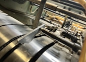 A close-up view of industrial machinery, featuring metallic cylindrical rollers and mechanical components interconnected with pipes and wires. The perspective highlights the mechanical complexity and precision engineering.