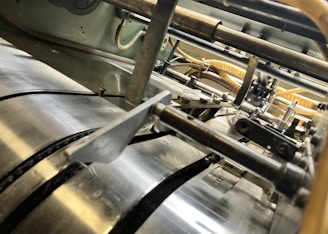 A close-up view of industrial machinery, featuring metallic cylindrical rollers and mechanical components interconnected with pipes and wires. The perspective highlights the mechanical complexity and precision engineering.