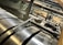 A close-up view of industrial machinery, featuring metallic cylindrical rollers and mechanical components interconnected with pipes and wires. The perspective highlights the mechanical complexity and precision engineering.