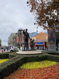 A statue of a man in a suit pointing forward, standing next to a famous animated character, is situated on a circular pedestal surrounded by a manicured garden with vibrant red and yellow flowers. The background reveals an amusement park setting, with a building entrance labeled 'Animation Celebration' and several people walking around.