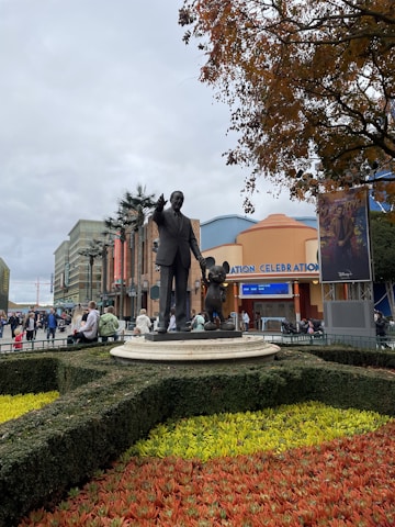 A statue of a man in a suit pointing forward, standing next to a famous animated character, is situated on a circular pedestal surrounded by a manicured garden with vibrant red and yellow flowers. The background reveals an amusement park setting, with a building entrance labeled 'Animation Celebration' and several people walking around.