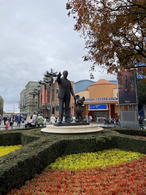 A statue of a man in a suit pointing forward, standing next to a famous animated character, is situated on a circular pedestal surrounded by a manicured garden with vibrant red and yellow flowers. The background reveals an amusement park setting, with a building entrance labeled 'Animation Celebration' and several people walking around.