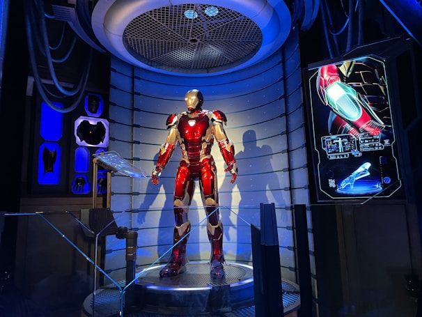 A high-tech suit displayed in a futuristic setting, illuminated by focused lighting. The surroundings include digital screens showing technical data and designs related to the suit. The environment appears to be a lab or exhibit with metallic and technological elements.