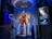 A high-tech suit displayed in a futuristic setting, illuminated by focused lighting. The surroundings include digital screens showing technical data and designs related to the suit. The environment appears to be a lab or exhibit with metallic and technological elements.