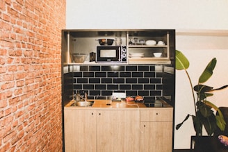 A compact kitchen setup integrated into a wooden cabinet with black tiled walls. There is a microwave oven, several kitchen utensils, plates, and bowls placed on shelves. The countertop includes a sink, a small electric stove, and some food items. A plant with large leaves is situated beside the kitchen against a white wall. The adjacent wall is covered with red bricks, adding a rustic touch to the setting.
