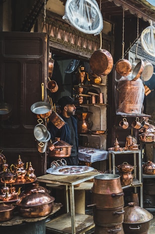 A shop displaying a variety of hanging and shelved copper kitchenware. The display includes pots, pans, and kettles with a man positioned next to the shelves. The scene showcases a traditional market atmosphere.