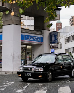 A black taxi car is turning at a street corner in front of a Lawson convenience store. The store is located beneath a concrete structure, and various signs and posters are visible in the window. The setting appears urban, with a mix of modern and older buildings in the background. Green leaves from nearby trees are partially in view at the top of the image.