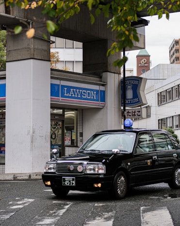 A black taxi car is turning at a street corner in front of a Lawson convenience store. The store is located beneath a concrete structure, and various signs and posters are visible in the window. The setting appears urban, with a mix of modern and older buildings in the background. Green leaves from nearby trees are partially in view at the top of the image.