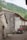 A rustic stone building with a red-tiled roof is situated in a mountainous, green area. A sign on the building reads 'Albergue Peña Main'. An off-road vehicle or quad bike is parked nearby. The surroundings include cobblestone paths and lush greenery, evoking a tranquil, rural atmosphere.