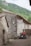 A rustic stone building with a red-tiled roof is situated in a mountainous, green area. A sign on the building reads 'Albergue Peña Main'. An off-road vehicle or quad bike is parked nearby. The surroundings include cobblestone paths and lush greenery, evoking a tranquil, rural atmosphere.